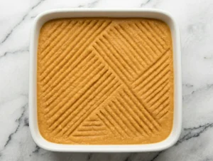 Smooth rutabaga mixture poured into a white square baking dish with a decorative crosshatch fork pattern pressed into the surface, before the breadcrumb and butter topping is added for Finnish lanttulaatikko