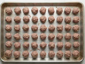 Overhead view of rows of small raw Swedish meatballs evenly spaced on a metal baking tray, ready to be refrigerated before frying