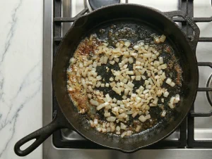 Top-down view of finely diced onion softening in butter in a well-seasoned cast iron skillet on a gas stove