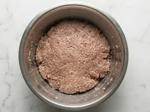 Overhead view of combined Swedish meatball mixture of ground beef and pork with breadcrumbs and onion in a stainless steel mixing bowl on a marble surface