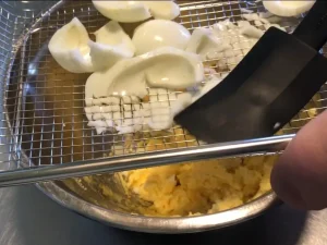 Close-up of hard-boiled egg whites being pressed through a wire cooling rack with a black spatula into a metal bowl to create uniform pieces for Munavoi.
