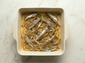 Top-down view of a baking dish layered with matchstick potatoes, sautéed yellow onions, and Swedish sprat fillets.
