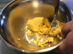 A stainless steel bowl containing a block of softened yellow butter and several hard-boiled egg yolks being mashed together with a fork.