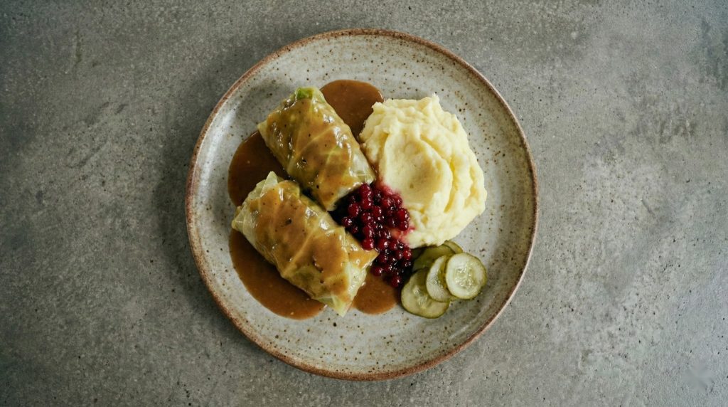 Swedish cabbage rolls (kåldomar) with brown gravy, creamy mashed potatoes, lingonberries, and pickles on a rustic plate.