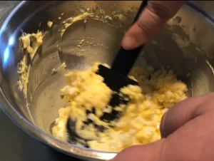 A person using a black spatula to gently fold diced egg whites into a thick, creamy yellow yolk and butter mixture in a stainless steel bowl.
