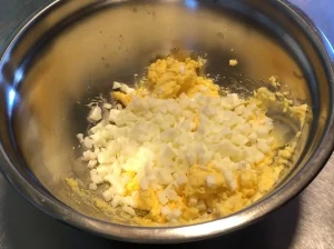 A pile of uniform, finely diced hard-boiled egg whites sitting on top of a creamy yellow egg yolk and butter mixture in a stainless steel bowl.
