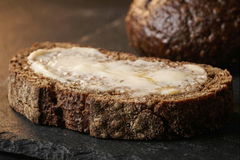 lose-up macro shot of a dense, dark slice of Finnish sourdough rye bread generously topped with a thick layer of yellow butter.