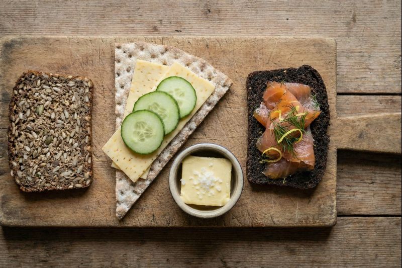 A wooden serving board arranged with three types of rye: a seeded Danish slice, a Swedish crispbread with cheese, and a sweet dark archipelago slice with salmon.