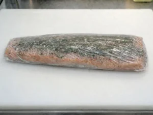 The seasoned and herbed salmon fillet tightly wrapped in multiple layers of clear plastic cling film.