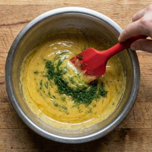 Folding chopped fresh dill into creamy Swedish hovmästarsås in a mixing bowl with a spatula.