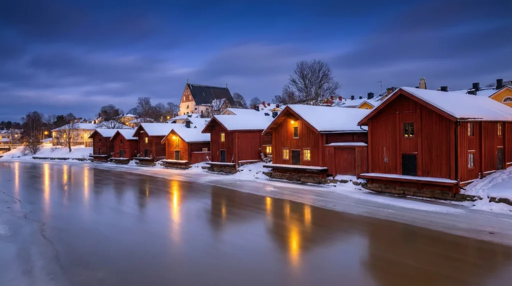 A panoramic view of Old Porvoo, Finland, at twilight. Iconic red ochre wooden warehouses line the frozen Porvoo River covered in snow, with the silhouette of the Porvoo Cathedral visible on the hill in the background.