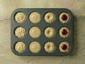 A 12-cup muffin tin partially filled with cake batter, showing small thumbprint indentations being filled with red raspberry jam.