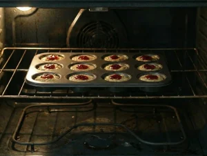 A muffin tin of Runebergintorttu cakes baking on the center rack of a lit oven.