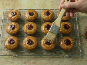 A hand using a pastry brush to apply a clear rum and sugar syrup to the tops of baked cakes on a wire cooling rack.
