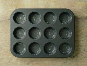 A top-down view of a 12-cup metal muffin tin on a wooden surface, with each cup visibly greased with butter.