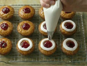 A piping bag applying a thick ring of white powdered sugar icing around the red jam center of a baked cake.