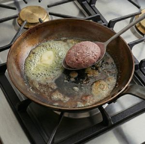 Shaped frikadelle resting on a spoon above a buttered pan, ready to be added for pan-frying.