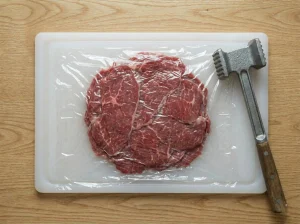 Beef tenderloin pounded paper-thin under plastic wrap on a cutting board next to a meat mallet, for Toast Pelle Janzon