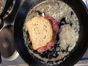 A slice of bread with beef on the bottom frying in a cast iron skillet with foaming butter.
