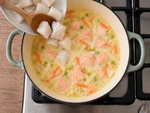 Chunks of cod being added to creamy Norwegian fish soup with salmon and vegetables.